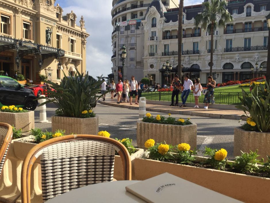 A view of Cafe de Paris, Monaco. In the foreground is an empty chair, in the background is the entrance to Monte Carol casino with tourists milling around.